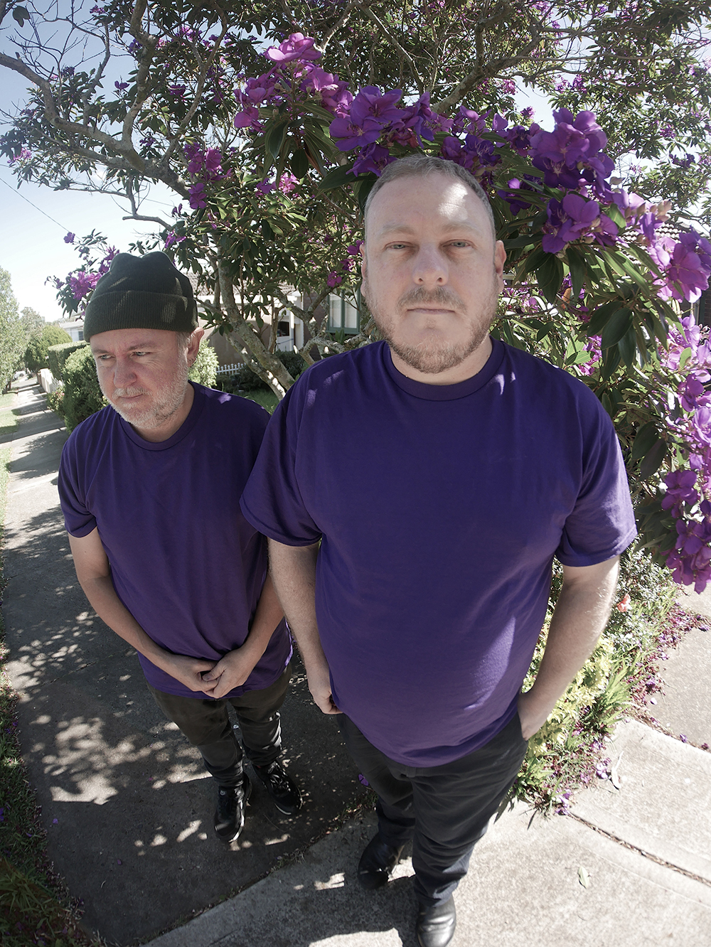 Two men in blue shirts and black jeans in front of a flowering vine. 