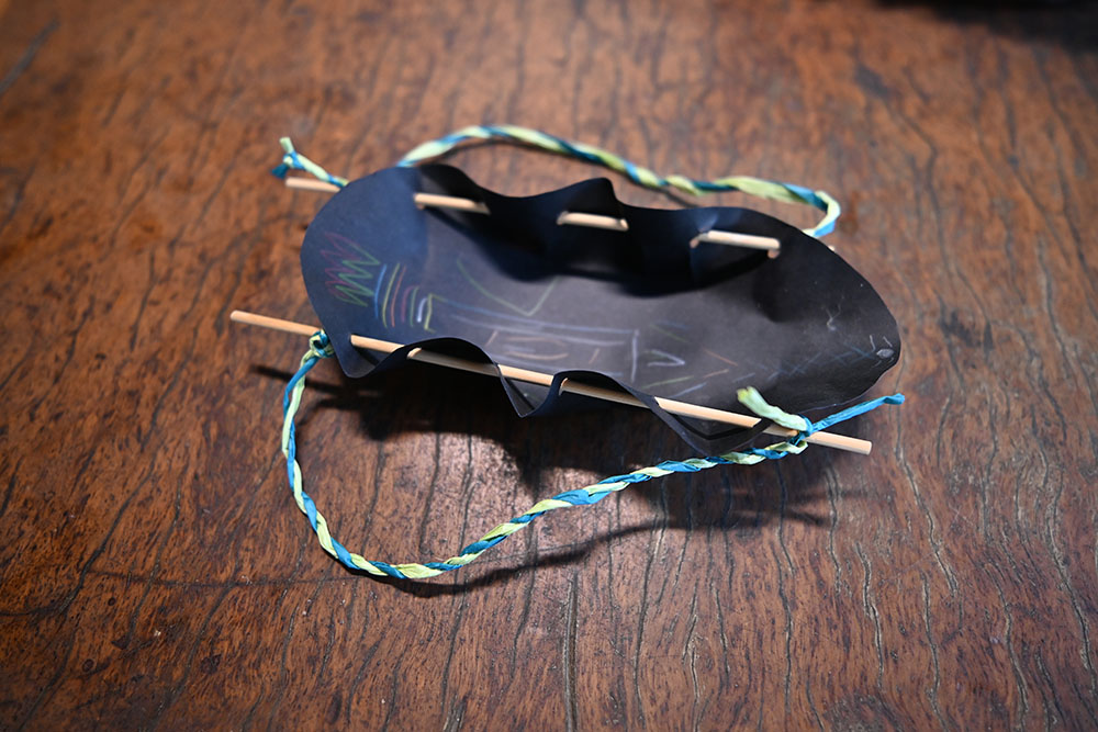 A photo of a handmade treasure carrier artwork. It is made from black carboard, string and wood. 