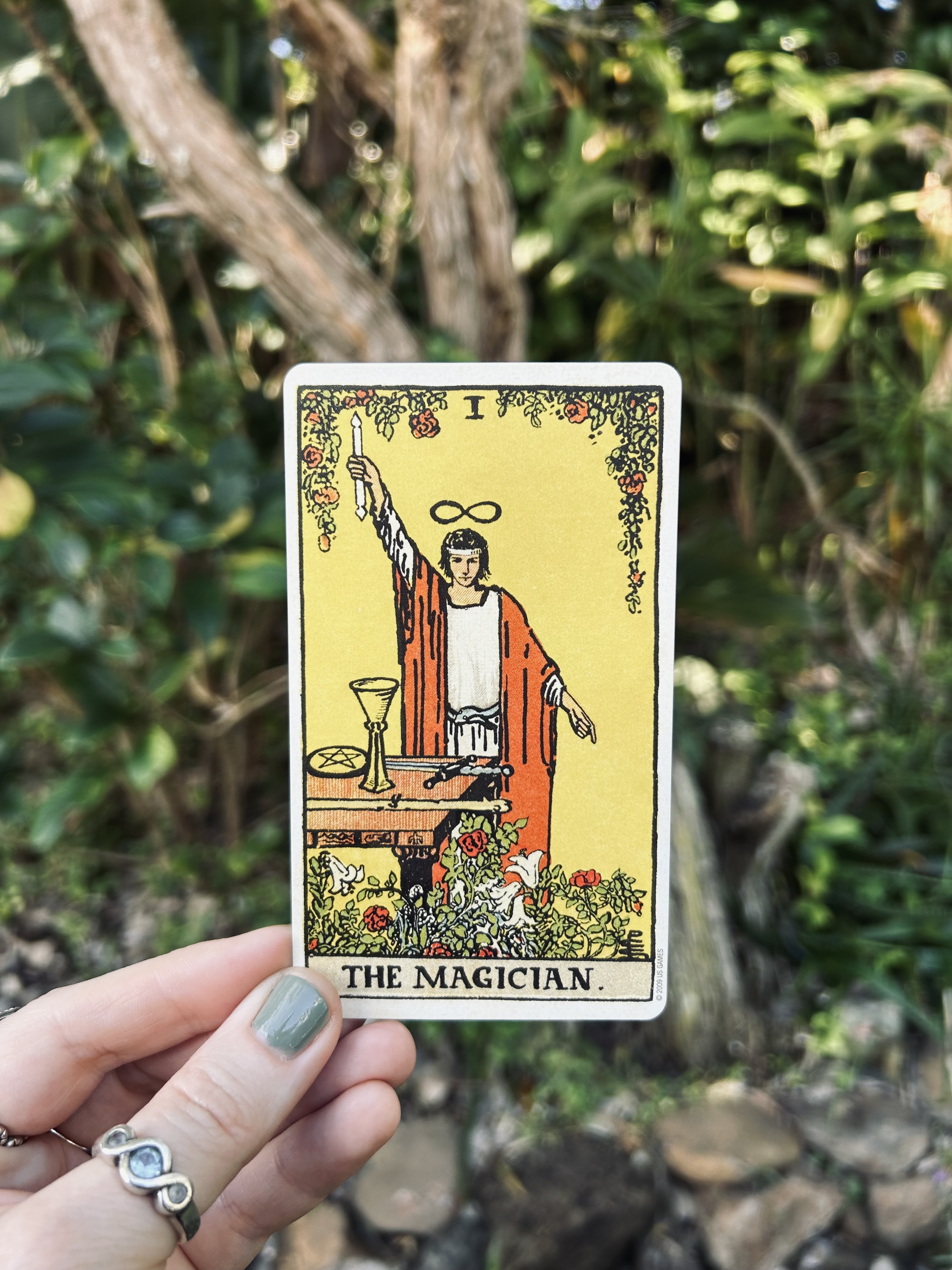 A photo of a hand holding The Magian Tarot card from the Smith Ride Waite deck. There is a garden in the background. 