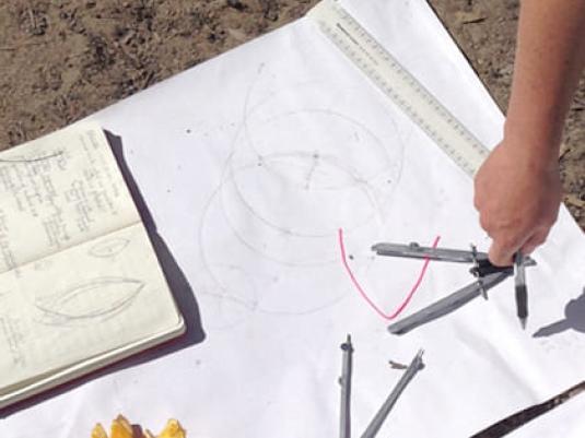 person using compass to draw curved line on paper. Open book, a ruler and orange peel is also in image