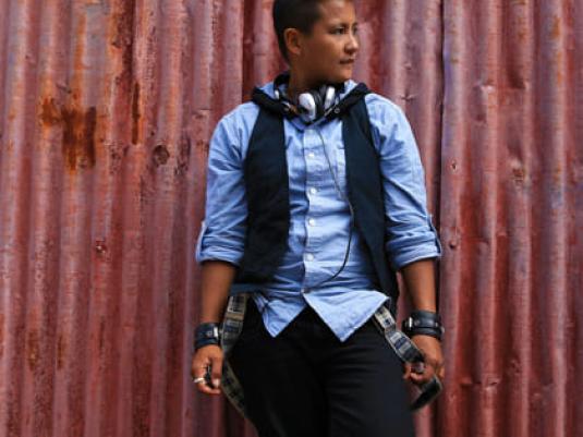 person in blue shirt with dark vest and trousers standing in front of corrugated iron shed