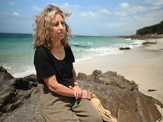 A photo of a woman with greying curly hair, a black t-shirt and brown pants. She is sitting on a rock with a stunning beach behind her