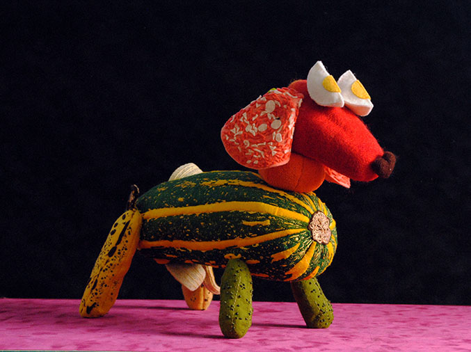 A soft sculpture sausage dog made from felt - the eyes are eggs, the body a melon, the ears sliced meat, front legs are gerkins, and the back legs are banannas 