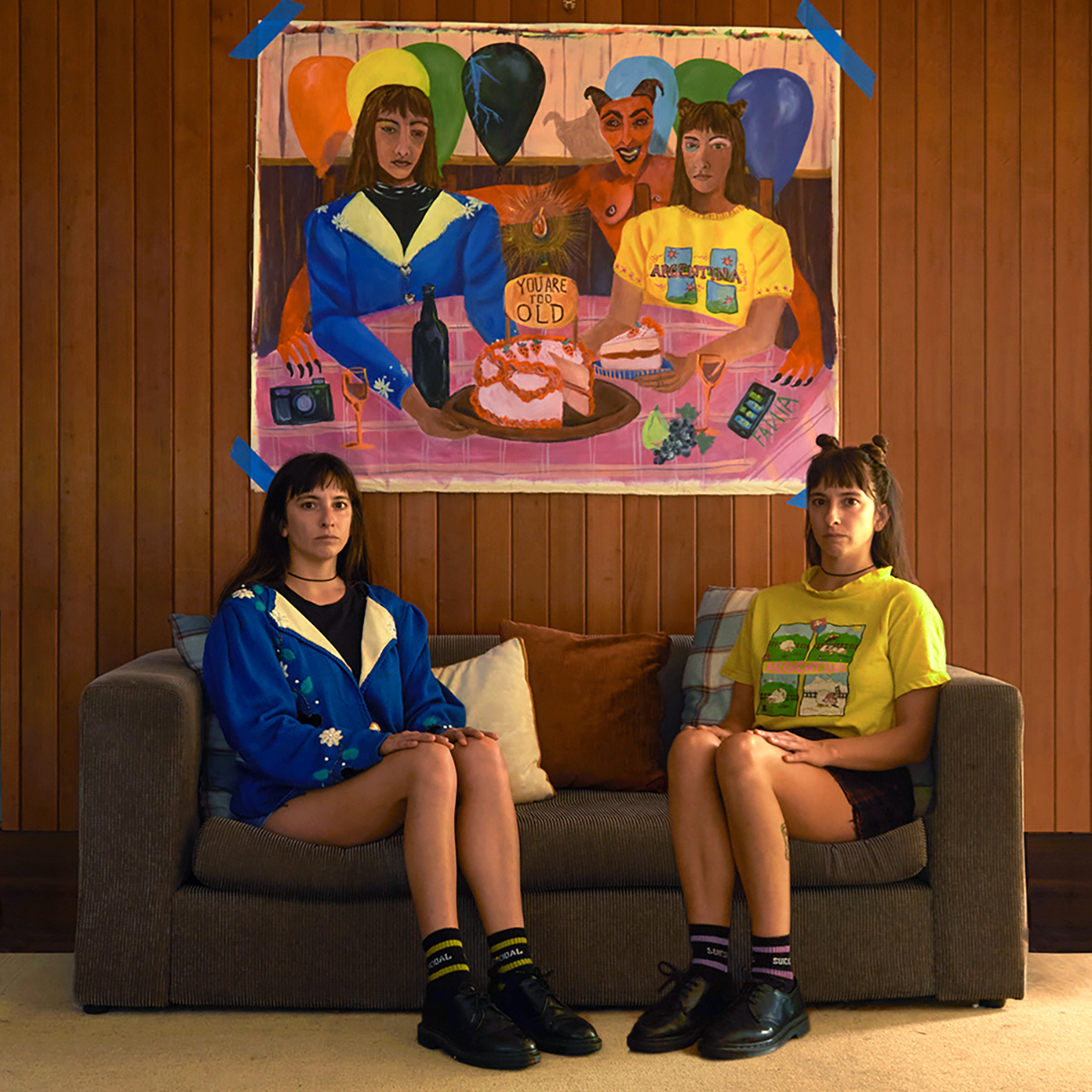 The artist in double, seated on two sides of the couch, with a self-portrait painting hanging above, echoing the double self below. 