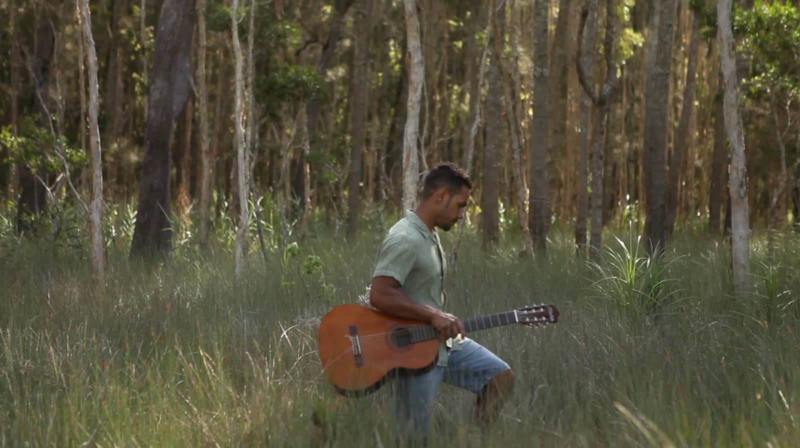 A young Aboriginal man walking through the bush, holding his guitar