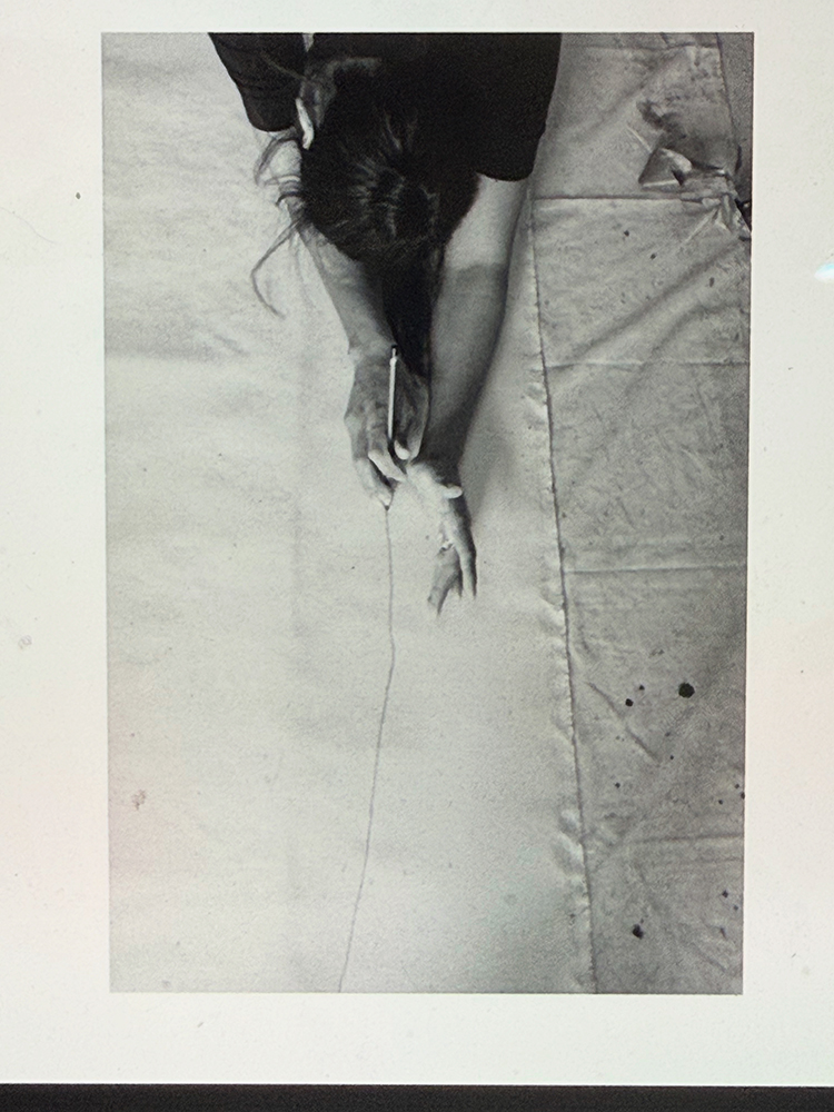 A black and white photo of a woman lying on a large sheet of canvas, marking it with a pencil
