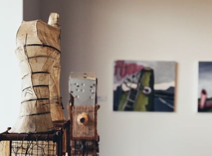 Sculptures made from found objects including some bed base wire, a pot, and box forms with a cylindrical sculpture that resembles a mannequin made from wire and paper in foreground with paintings on wall baind 