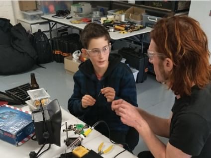 Two people ollaborating on creating an object. There is a desk lamp, soldering iron, pliers and other tools and equipment on the table they are sitting at, and on the table in the background