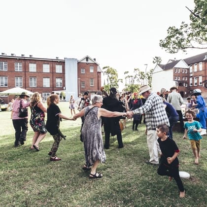 group of people in the gallery Quad. Some children running around, spome people holding hands and dancing in a circle with other  people are gathered in background e 