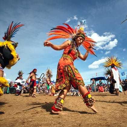 woman in Sioux tribal dress with headdress dancing in foreground with other people dancing in background in tribal dress.