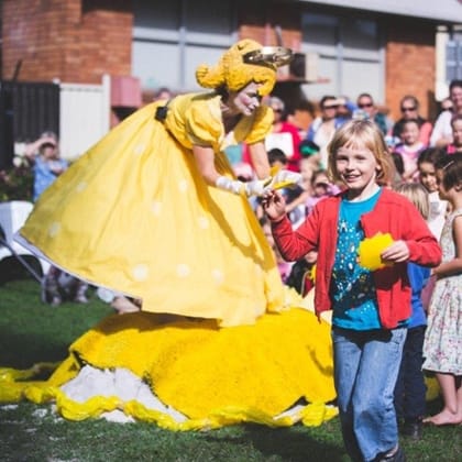 a crowd of children behind a woman dressed in yellow frock with wide skirt with a yellow headdress giving a child somethin yelloww