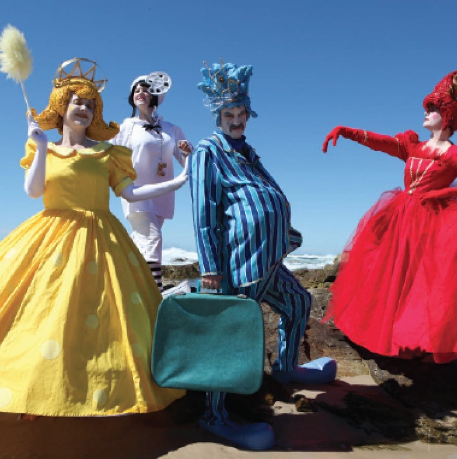groups of people dressed in comedic costume - a woman in yellow hooped dress with gold hair carrying a feather duster, a woman in red dress and headdress, a man wearing blue striped outfit, oversized shoes and a blue hesddress with a woman behind them all dressed in white outfit. The ocean and bright bule sky are behind them all.  