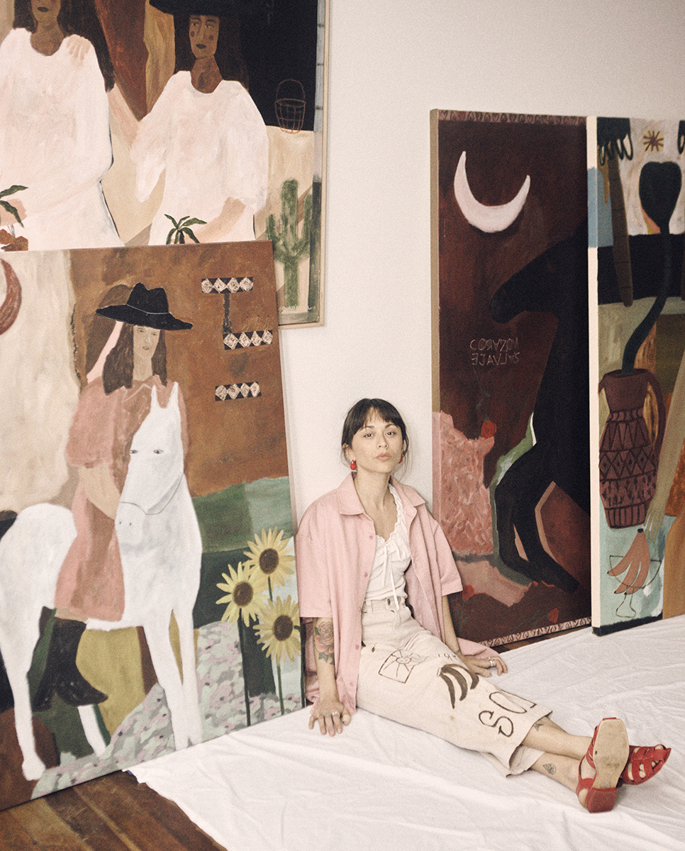 A Chilean/ Argentine female painter sitting next to her large canvases. The imagery is brown and warm hues, with women, horses and moons