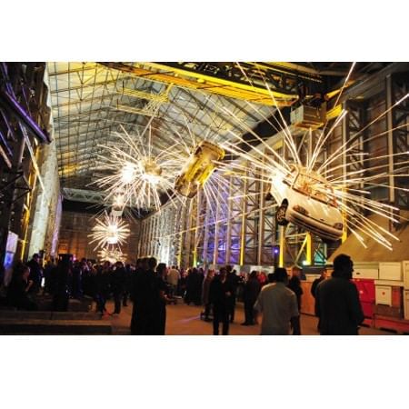 crowd of people below artwork that consists of cars suspended  from high ceiling with light rays coming out of them s coming 