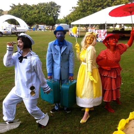 people dressed in the primary colours of re, blue and yellow, with a person in white  in from of a large tent in the background
