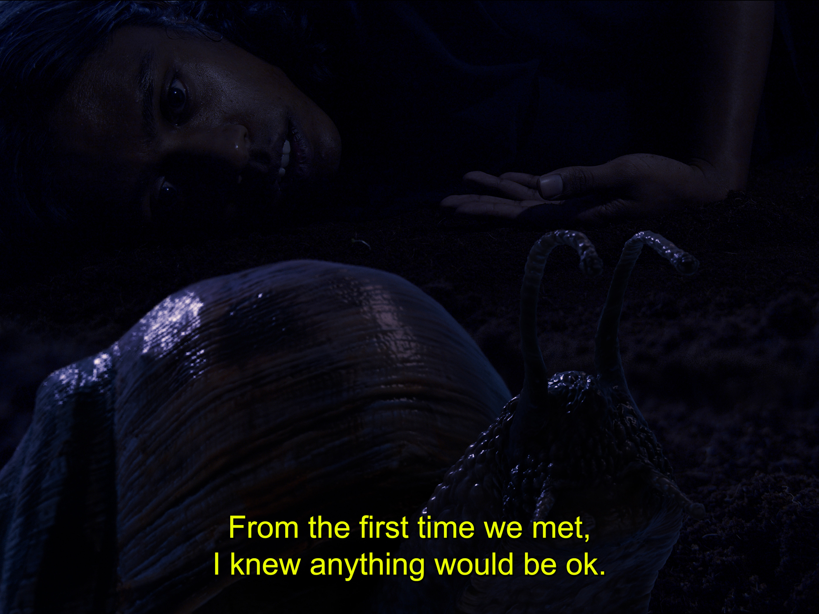 A film still featuring a dark, dimly lit frame, mostly black and purple with the silouette of a woman lying next to a large snail. The image has a subtitle saying ' From the first time we met, I knew anything would be ok'