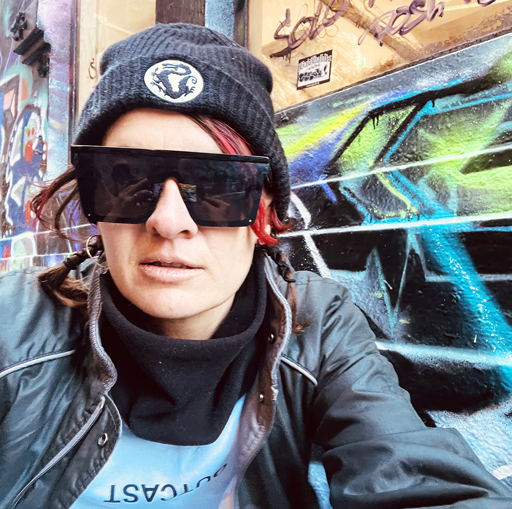 A portrait of the artist JAJA, they/them. They are wearing a black beanie and black sunglasses and sitting in front of a graffitied wall