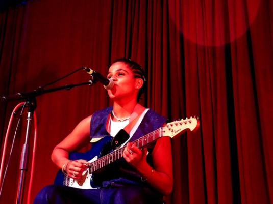 First Nations musician Angel White, pcitured on stage in front of a red velvet curtain, sitting on a chair, wearing a blue dress, playing guitar and singing