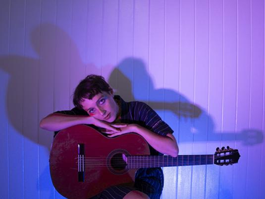 Musician Myki Jay, sitting with their guitar, in a room with a blue and purple hue