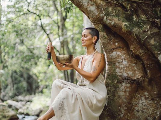 A woman of colour sitting on the root of a huge fig tree, holding a singing bowl instrument.There is a rainforest creek in the background.