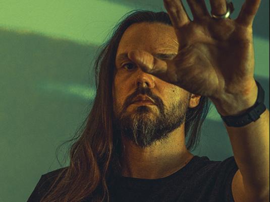 A man with long hair holds his open palm in front of his face and eye. He is wearing black and standing in front of a green background