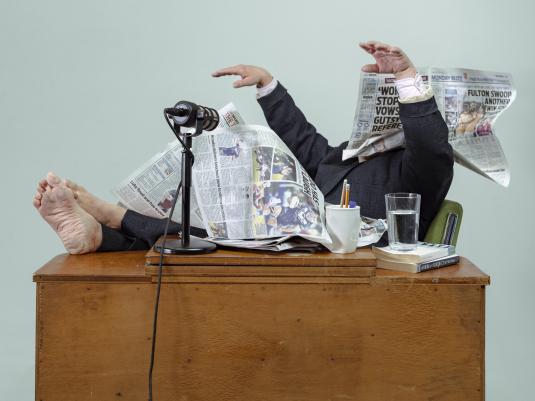 a person in a men's suit with feet on a timber desk, arms in the air and face and much of their body covered by newspaper pages.
