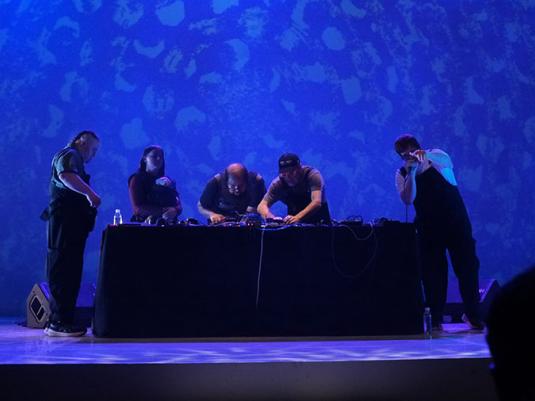 A group of musicians playing live on stage. They are standing around a table with electronic equipment with a blue backdrop