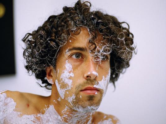 A portrait of a man with curly hair and white paint over his face and body