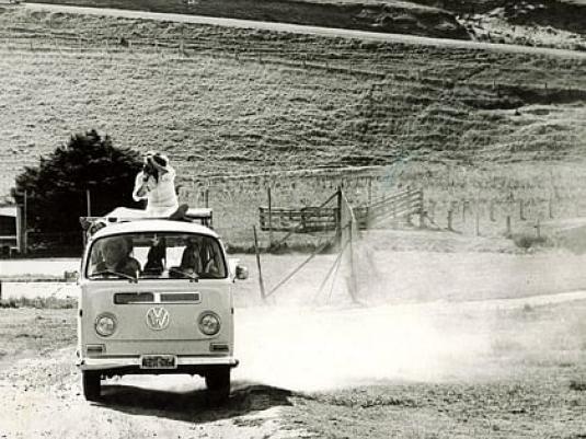 a kombi being driven on a dirt road with a photographer riding on the roof