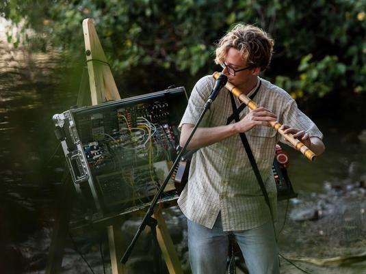 A young man playing a type of flute next to a river. There is a large electronic music making device as well.