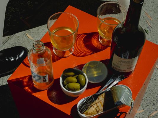 A photo of wine, olives ans fish, on a red table with the afternoon sun creating nice shadows