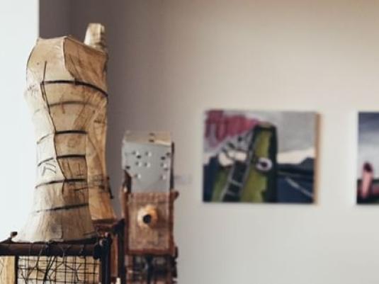 Sculptures made from found objects including some bed base wire, a pot, and box forms with a cylindrical sculpture that resembles a mannequin made from wire and paper in foreground with paintings on wall baind