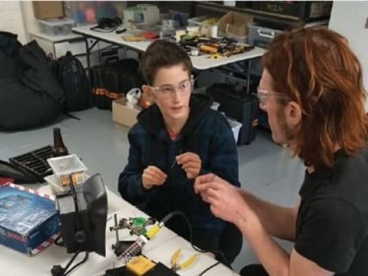 Two people ollaborating on creating an object. There is a desk lamp, soldering iron, pliers and other tools and equipment on the table they are sitting at, and on the table in the background