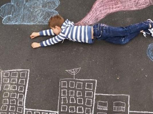 child lying on ground with drawings of building below them and a cloak drawn on ground so it appears he is wearing it and flying over the buildings and under the cloud