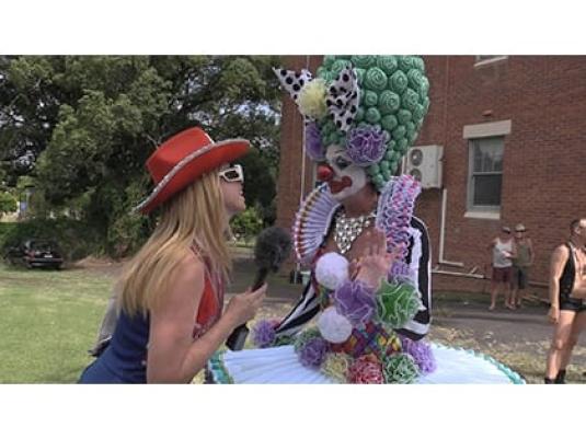 Maude Boate with painted clown face in elaborate wig and frock talking to Dee Dee Chenille who is wearing a red wide brimmed hat