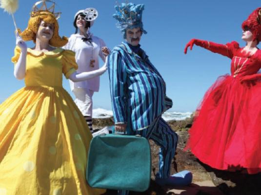 groups of people dressed in comedic costume - a woman in yellow hooped dress with gold hair carrying a feather duster, a woman in red dress and headdress, a man wearing blue striped outfit, oversized shoes and a blue hesddress with a woman behind them all dressed in white outfit. The ocean and bright bule sky are behind them all.