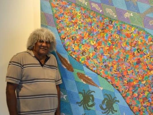 Digby Moran standing next to one of his paintings that includes a bright colourful island in the middle with fish and crabs swimming around it on a background of intricately patterned squares