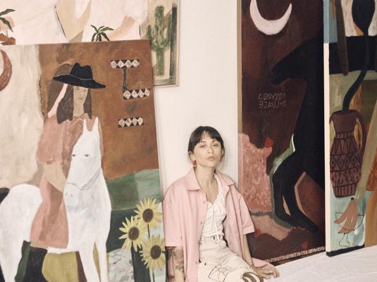 A Chilean/ Argentine female painter sitting next to her large canvases. The imagery is brown and warm hues, with women, horses and moons