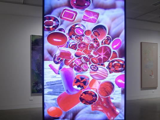 An exhibition documentation photo of a vertical screen showing a video with bright pink and purple jewel like objects floating