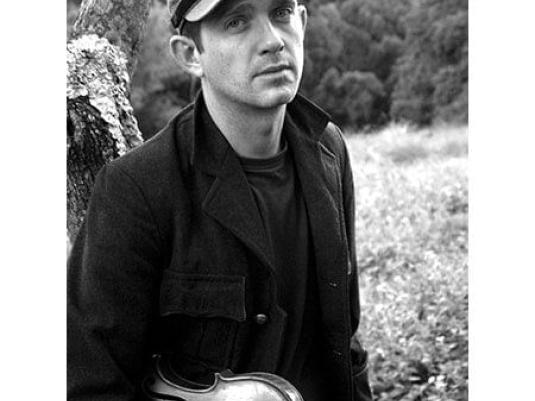 Cye Wood holding with a violin under his arm in front of a tree trunk, a  field and forest in background