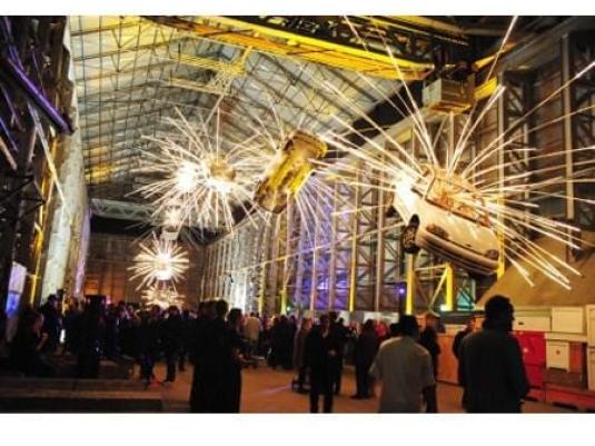 crowd of people below artwork that consists of cars suspended  from high ceiling with light rays coming out of them s coming