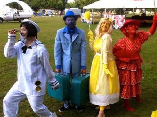 people dressed in the primary colours of re, blue and yellow, with a person in white  in from of a large tent in the background