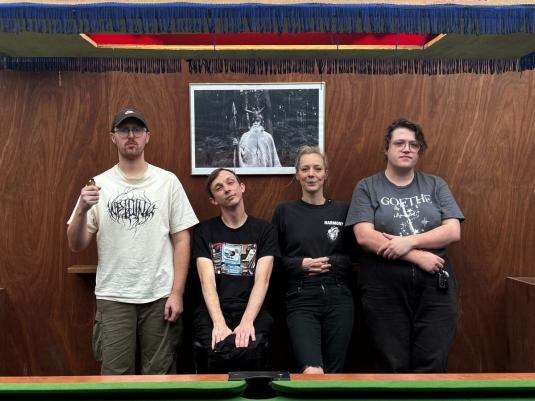 A punk band of four people, two males, one female, one non-binary, leaning against a wall in a pub, with a pool table in front of them.