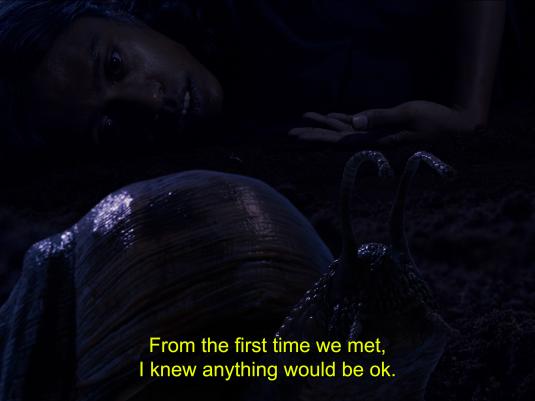 A film still featuring a dark, dimly lit frame, mostly black and purple with the silouette of a woman lying next to a large snail. The image has a subtitle saying ' From the first time we met, I knew anything would be ok'