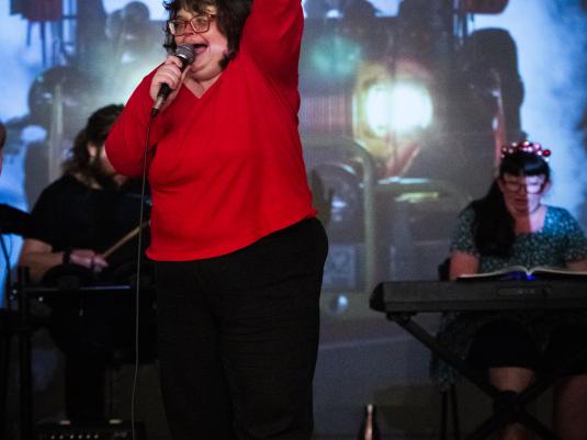 A woman with mixed abilities singing into a microphone with an arm raised in joy, in front of a projection