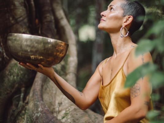 A woman of colour in a gold dress holding a singing bowl and looking up at the rainforest surrounding her