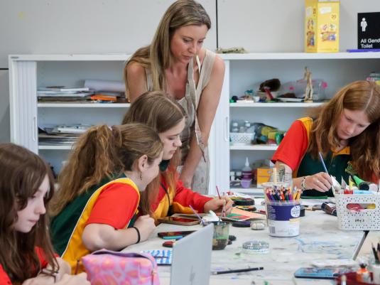4 female highschool students at a table making art, with their female teacher assisting them